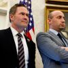 Двоє чоловіків стоять перед прапором США. Один складений руки.