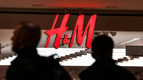 Getty Images яскраво-червоний логотип H&M, що відображається на знаку магазину, із силуетами двох перехожих проходять повз нього. 