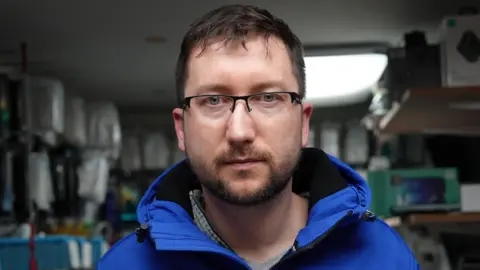 Себастьян Ліберек, дивлячись на камеру у своїй майстерні, в якій є телефони для продажу на задньому плані. Він носить синє пальто.